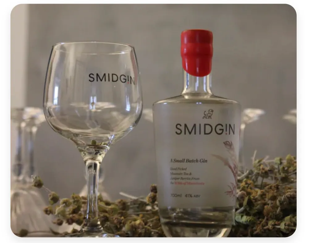 A branded Smidgin copa glass beside a bottle of Smidgin Classic on a bed of dried botanicals